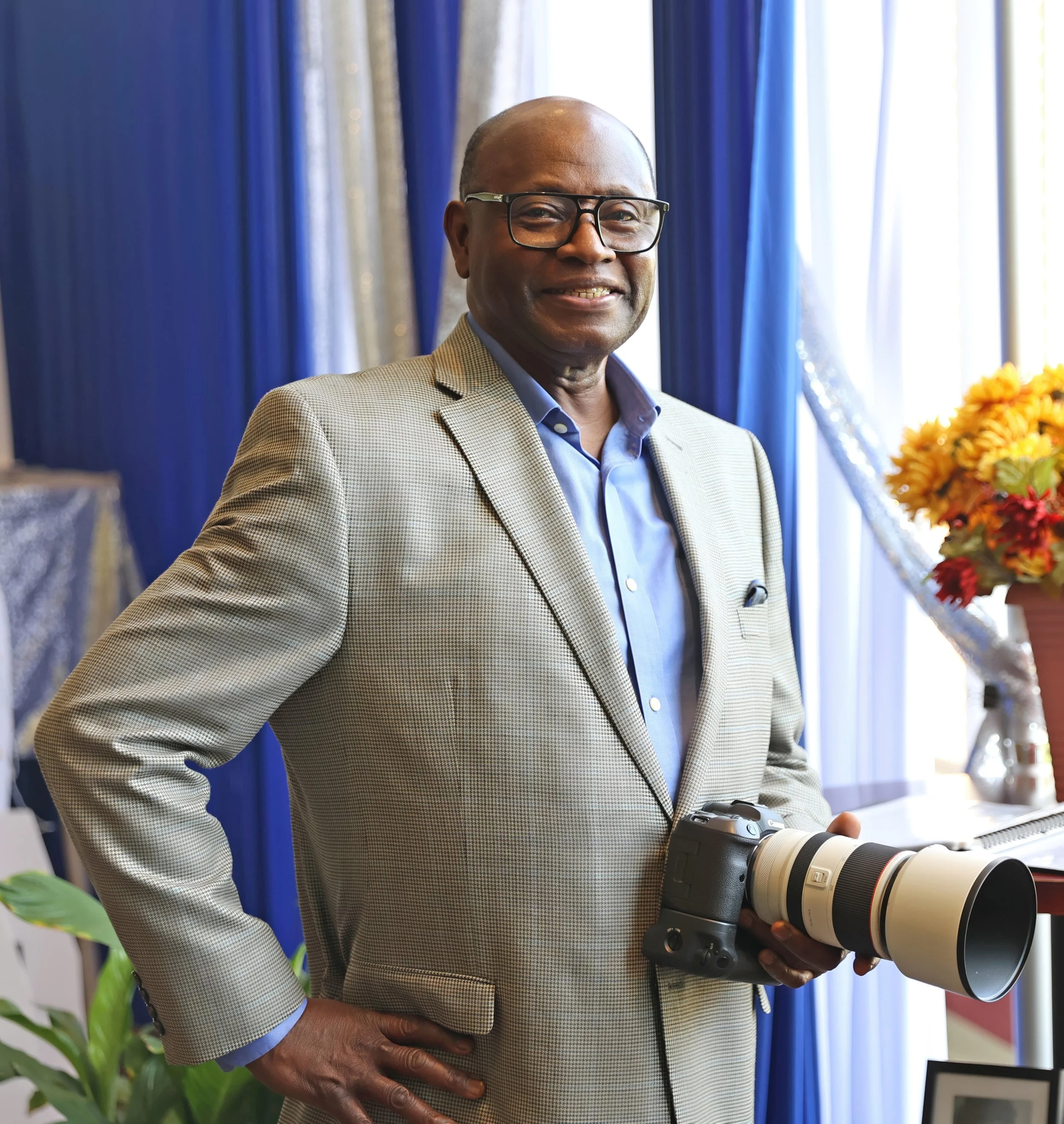 A man with glasses holding a large camera lens, standing indoors near blue curtains and a window with light shining through, with a bouquet of flowers on the side.