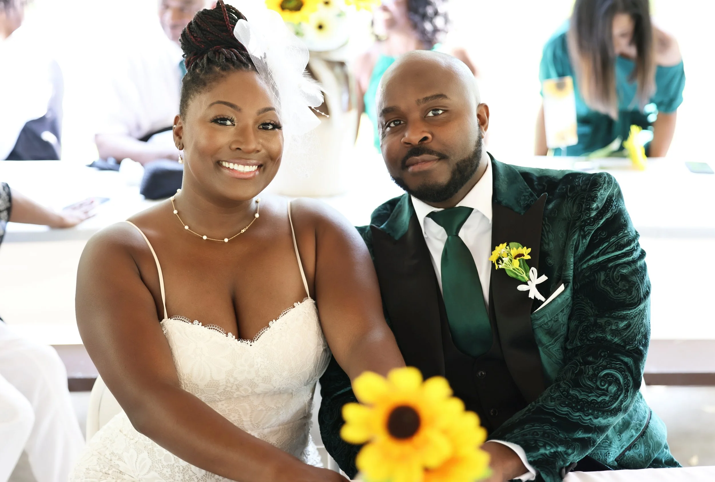 A smiling woman in a white lace dress and a man in a green suit with a white shirt and a green tie sitting together at a wedding reception. The woman has her hair styled in an updo with a white fascinator, and the man has a boutonniere of yellow flowers on his lapel. There are blurred people and flowers in the background.