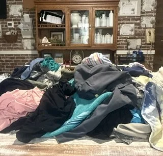 A cluttered table covered with various clothing items in front of a brick wall with a wooden hutch filled with dishes and a clock.