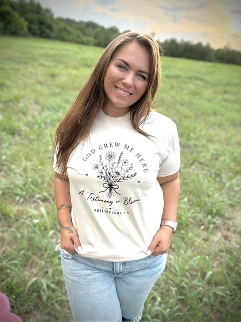 A young woman with long, light brown hair smiling outdoors in a grassy field, wearing a white T-shirt with floral design and text, and light blue jeans.