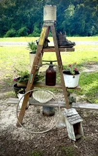 A rustic wooden ladder with gardening tools and potted plants, placed outdoors in a grassy area with trees in the background.