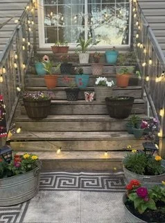 Flower pots with blooming flowers on stairs decorated with string lights, leading to a screened porch with a window and potted plants.