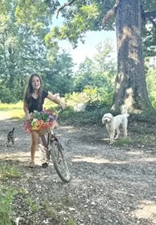 Young woman with long hair riding a bicycle with a basket of flowers on a dirt path in a park, accompanied by two dogs and a cat, surrounded by green trees.
