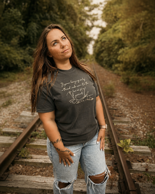 Young woman standing on train tracks surrounded by trees on a cloudy day, wearing a black t-shirt and ripped jeans.