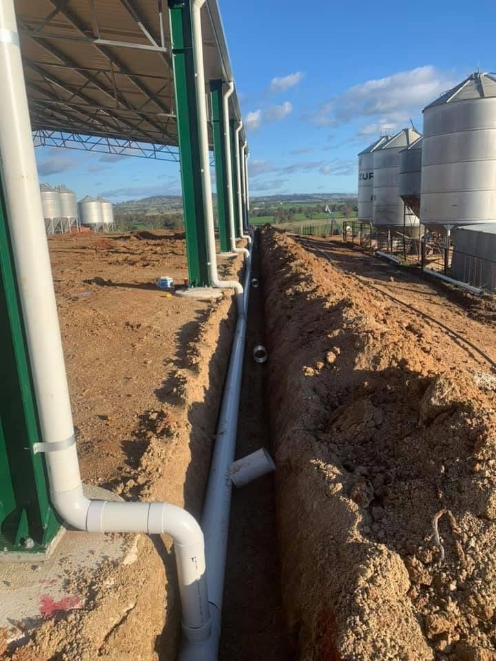 Pipes installed along a trench at an outdoor industrial site, with large metal tanks in the background and a partly cloudy sky.