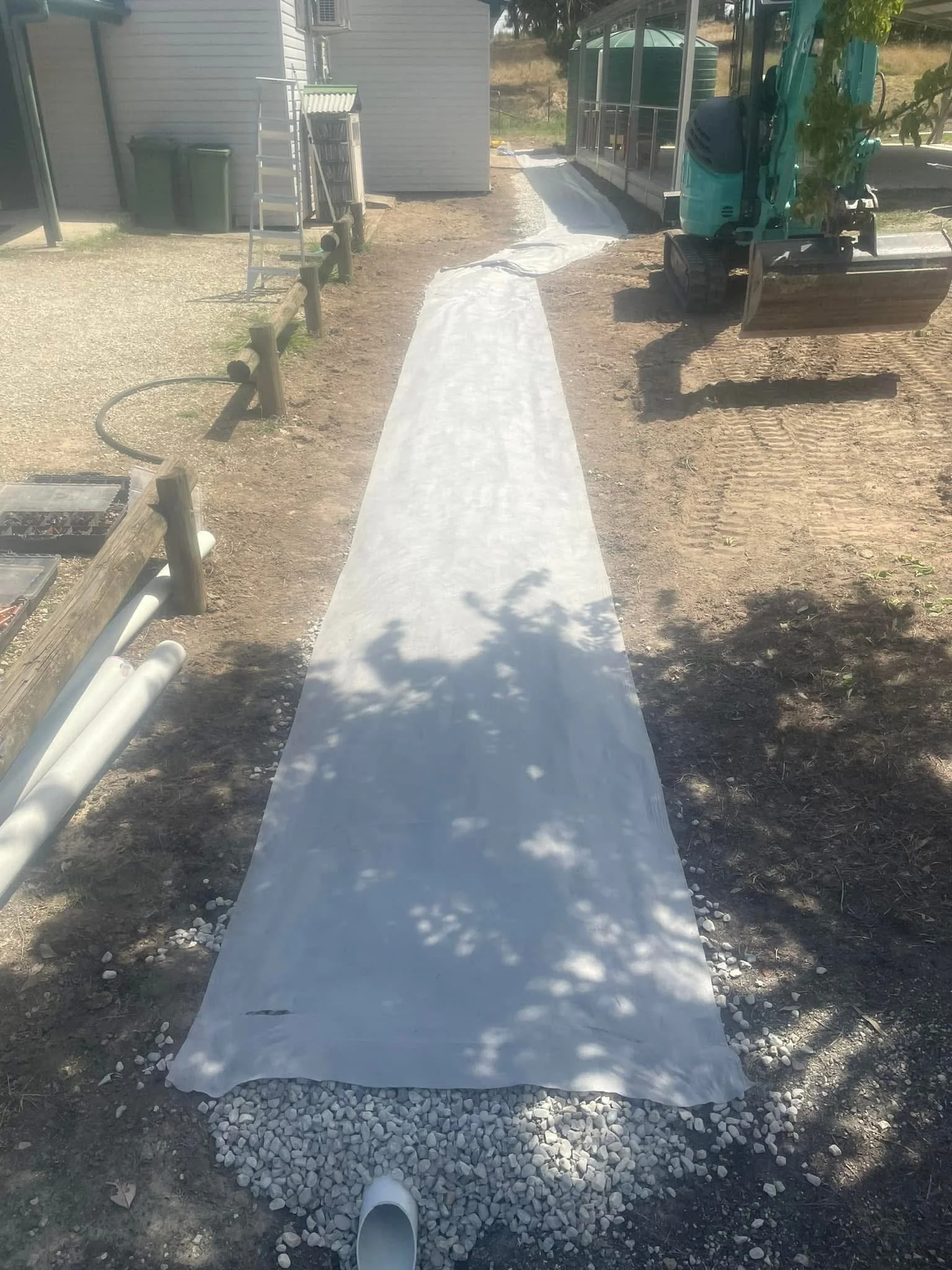 A concrete sidewalk with gravel at the edges, next to a hose and pipes, a ladder, and construction equipment near a house and a screened porch.