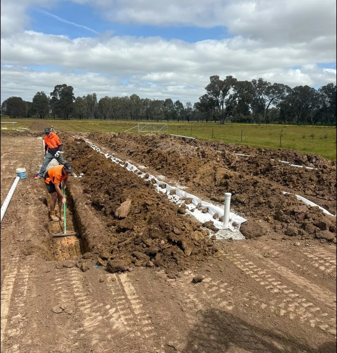 Two construction workers in orange shirts working on installing underground pipes in a field on a cloudy day. They are using tools to dig and place pipes along a trench with equipment and dirt surrounding them.