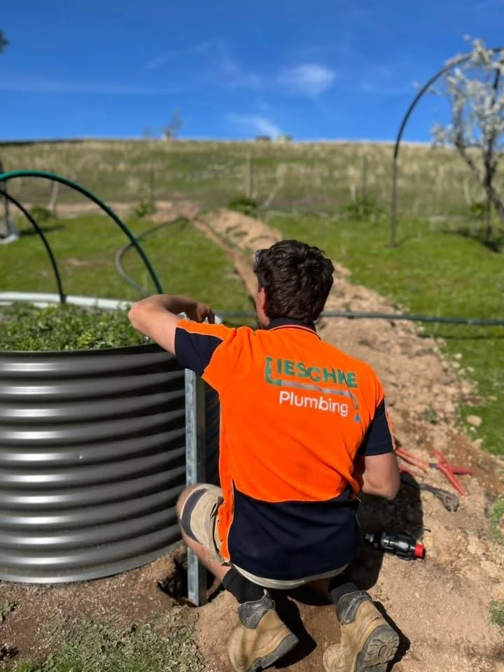 A man wearing an orange and black shirt with 'Liesch Plumbing' on the back, working outdoors on a plumbing or irrigation project near a raised garden bed with plants, in a grassy area under a blue sky.