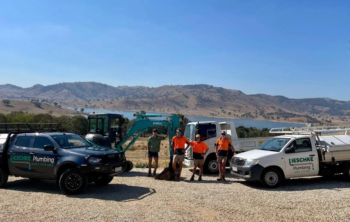Group of five plumbers standing in front of plumbing company vehicles outdoors on a gravel surface with hills and a lake in the background.