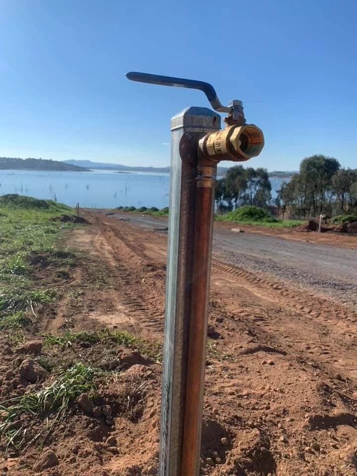 A fire hydrant with an upside-down spout located on a dirt road beside a body of water and some trees in the background.