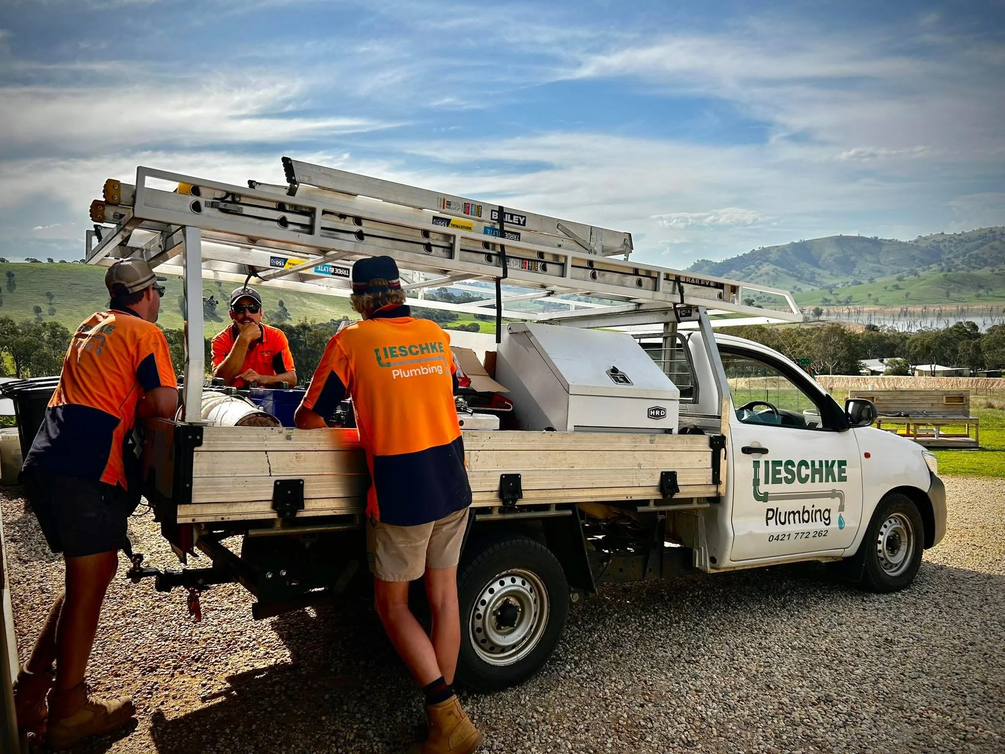 Three workers in orange shirts and one in a red shirt gathered around a white utility truck with a ladder mounted on top. The truck has company branding for "Lieschke Plumbing". They appear to be discussing something, with a scenic backdrop of hills 