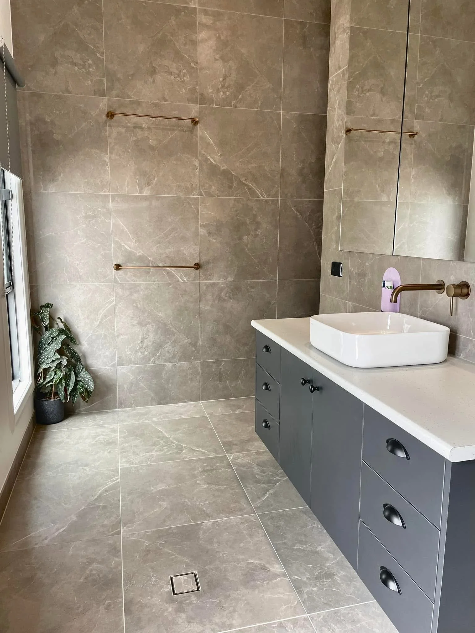 Modern bathroom with beige marble tiles on walls and floor, gray vanity with three round black handles, a white rectangular vessel sink, wall-mounted bronze faucets, a large mirror, two bronze towel bars on the wall, a small potted plant in a black p
