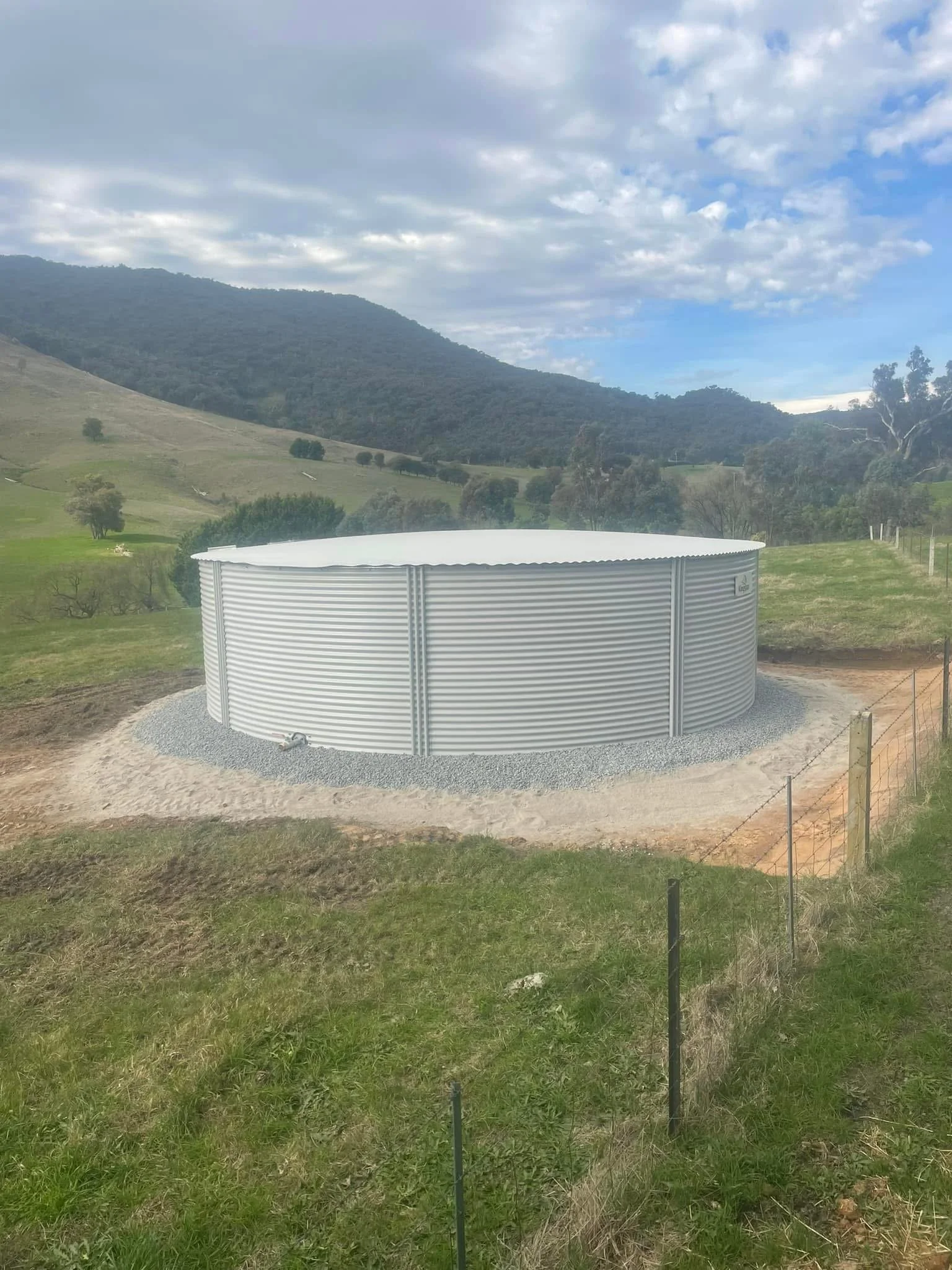 A large cylindrical water tank made of corrugated metal with a white curved roof is situated on a gravel base in a rural landscape. Surrounding the tank is green grass, a wire fence, and distant rolling hills with trees, under a cloudy sky.