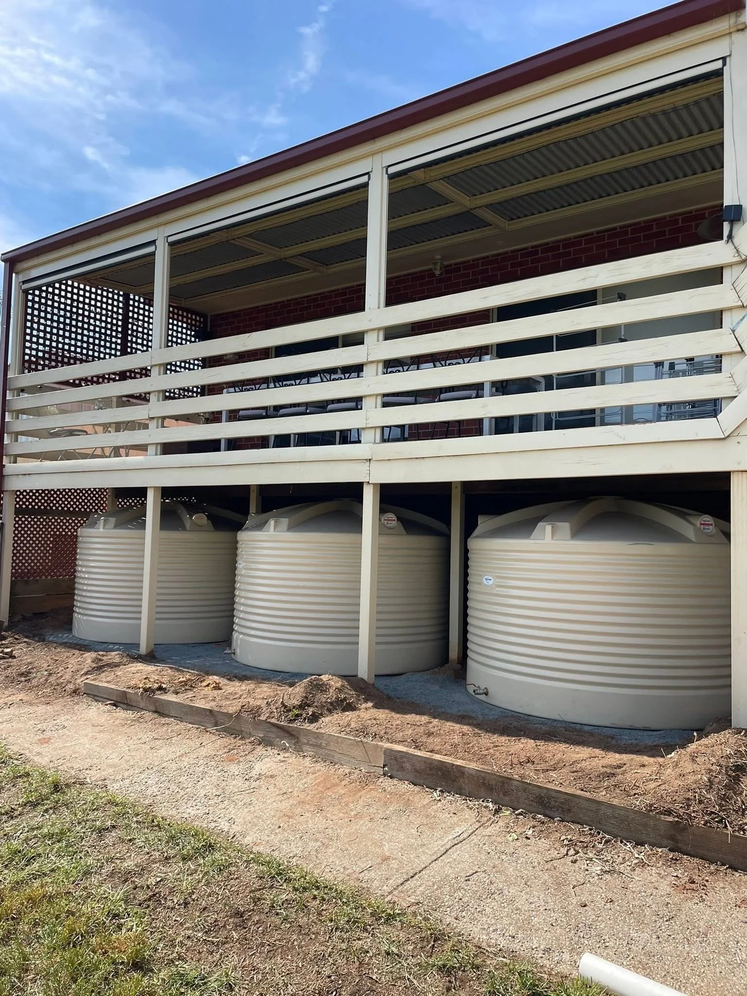 Three large white water tanks installed under a two-story house with a balcony. The house has a brick exterior and a red roof.