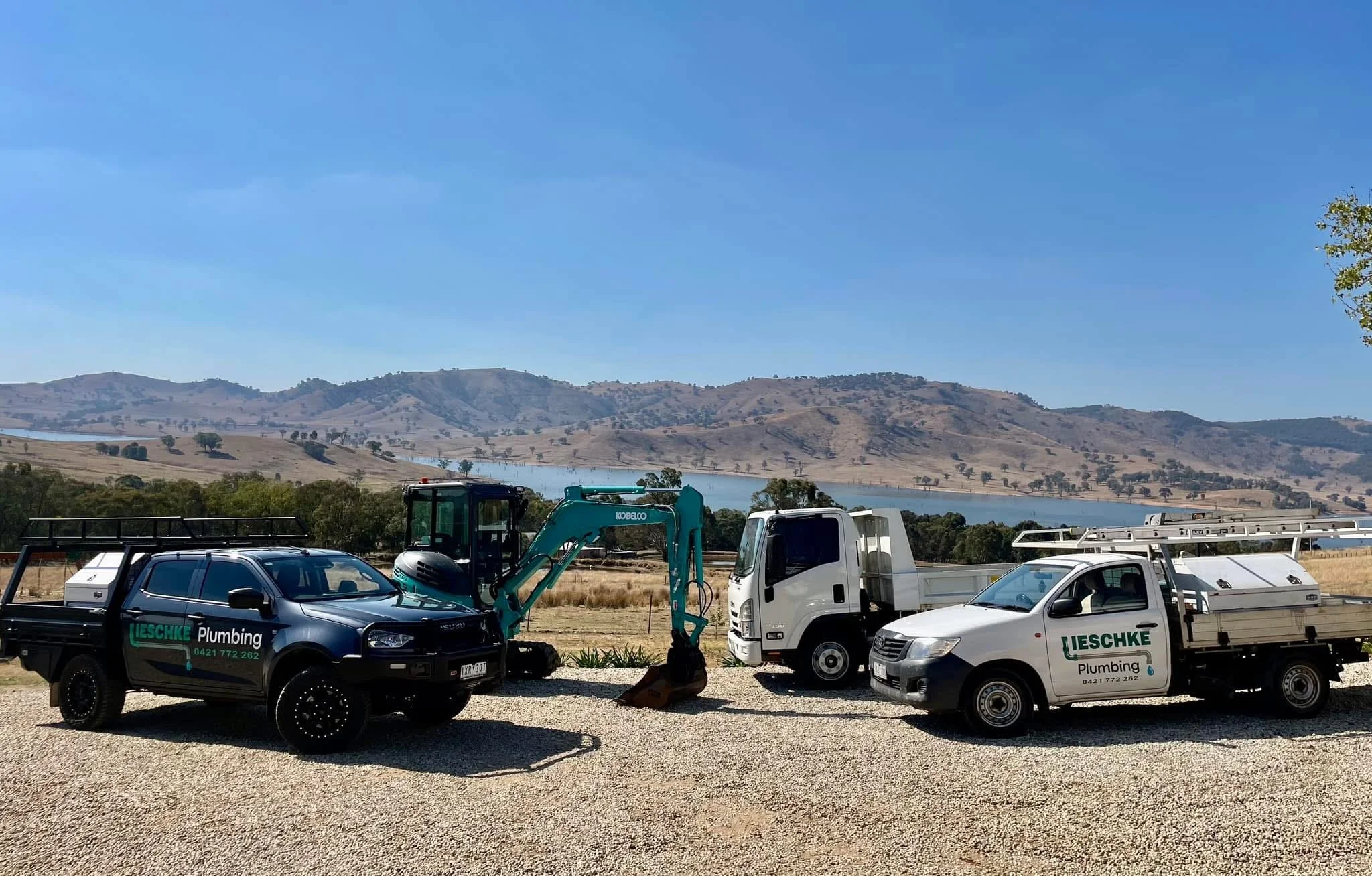 A construction lot with trucks and equipment, including a black truck and white truck with green company logos, set against a background of rolling hills and a lake under a clear blue sky.