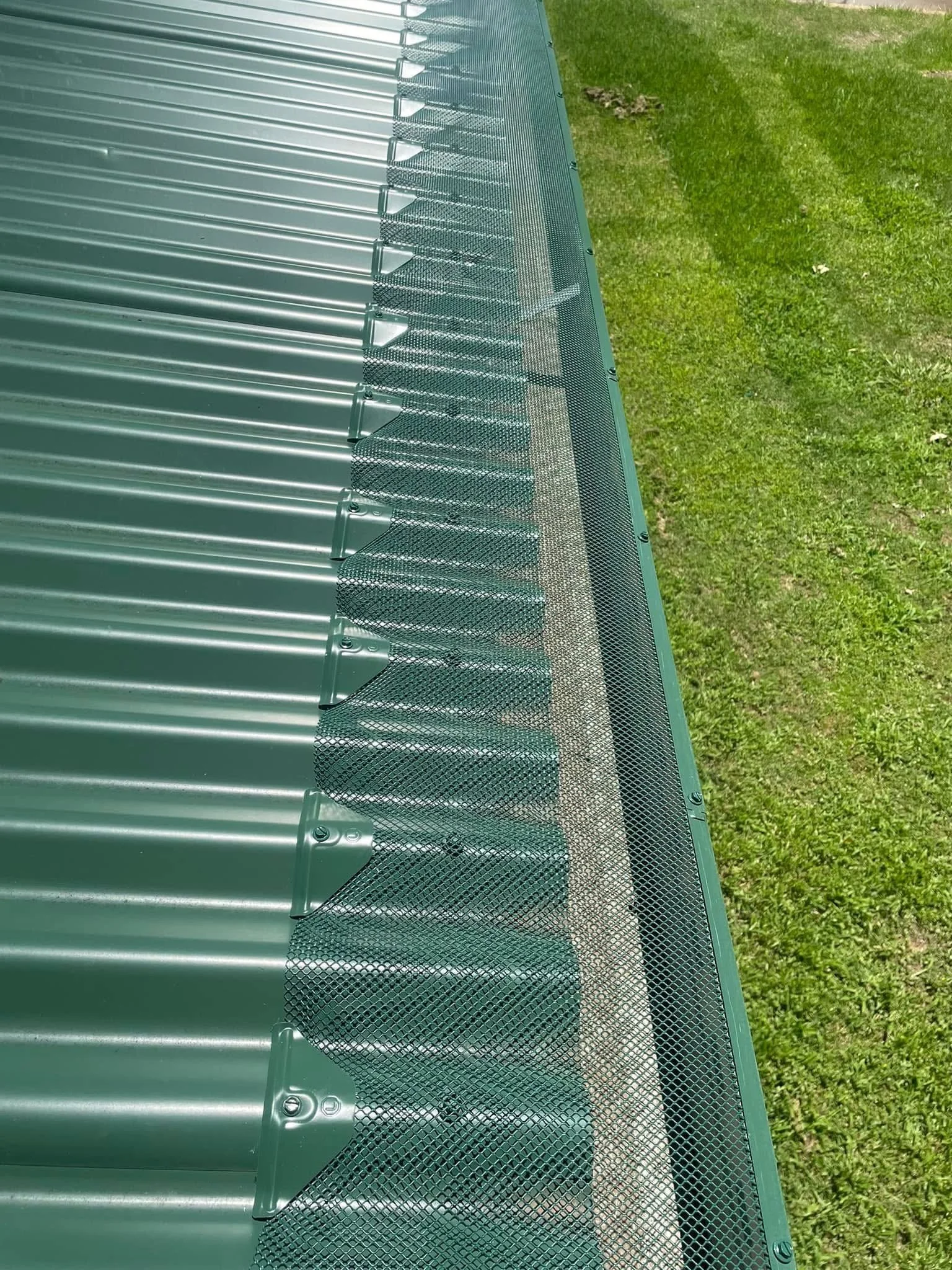Close-up view of the edge of a metal roof, showing metal ventilation screens along the top, with green grass in the background.