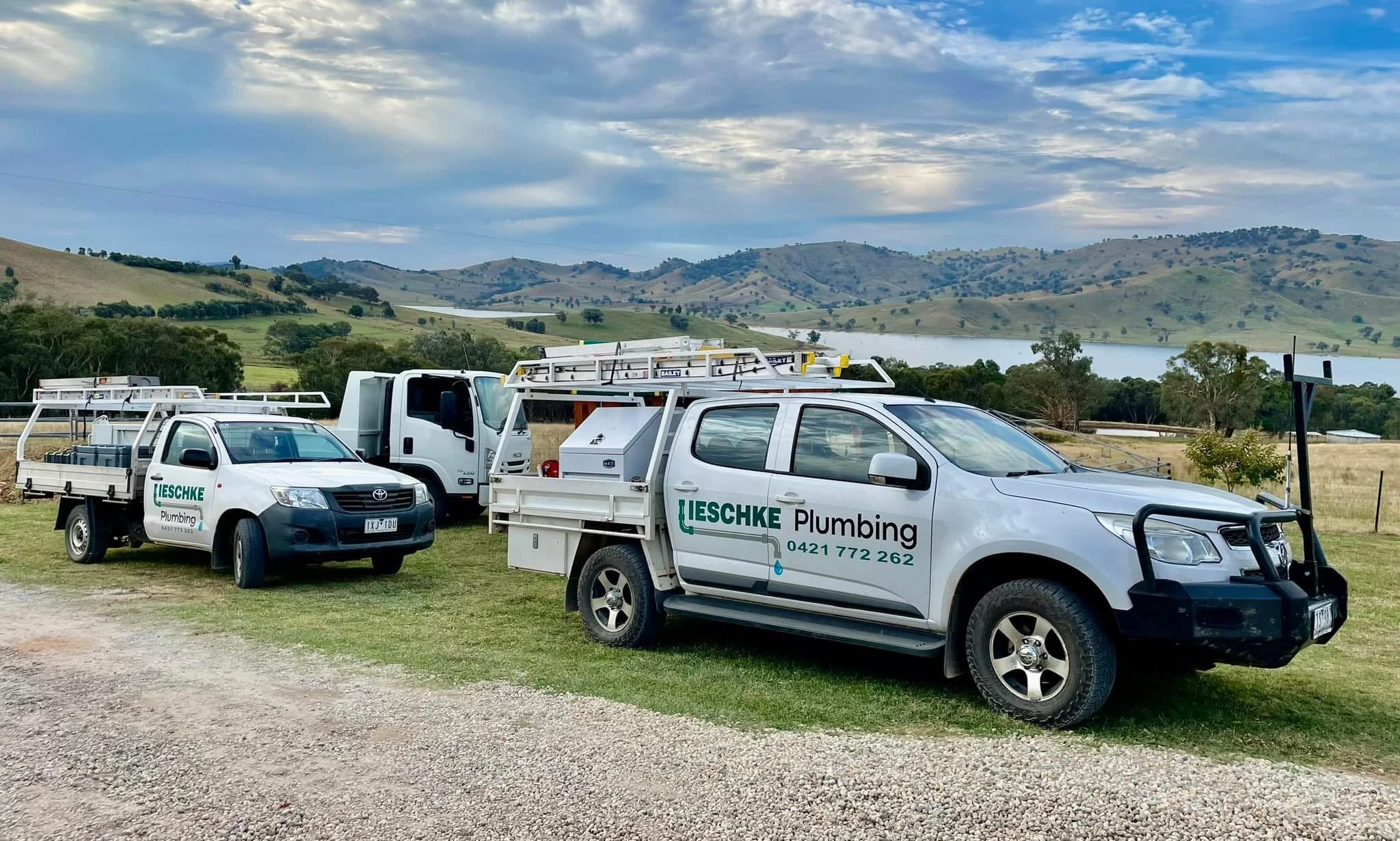 Three white service trucks with green and black lettering and equipment on top, parked on grass with rolling hills, trees, and a cloudy sky in the background.