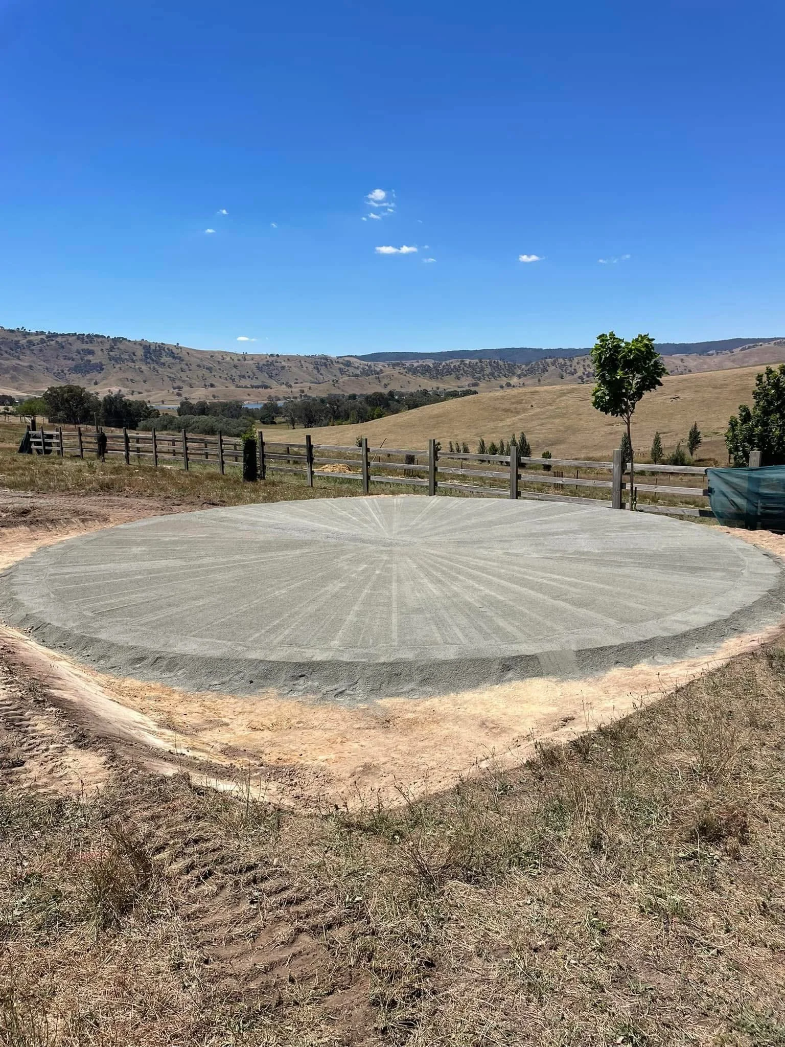 A circular area of freshly laid concrete in a rural setting with a wooden fence, grass, trees, hills, and a blue sky with scattered clouds.
