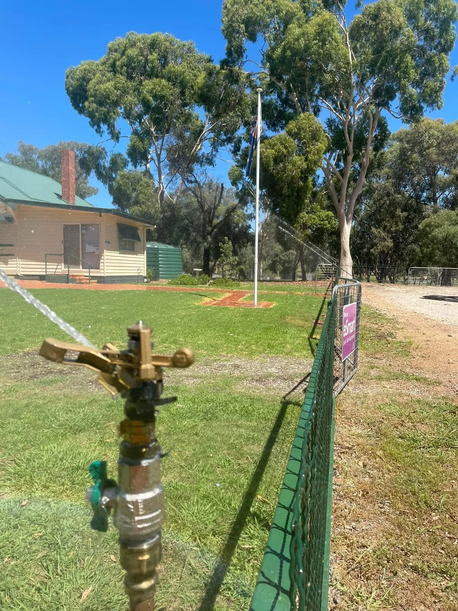 A fire hose connected to a water valve in a grassy area, with trees, a building, and a clear blue sky in the background. A green fence runs along the right side of the scene.