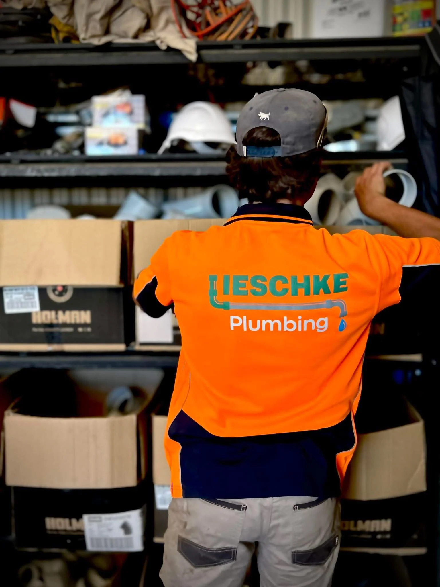 A Lieschke Plumbing worker in an orange branded shirt standing in front of shelves stocked with plumbing supplies and equipment in a warehouse.