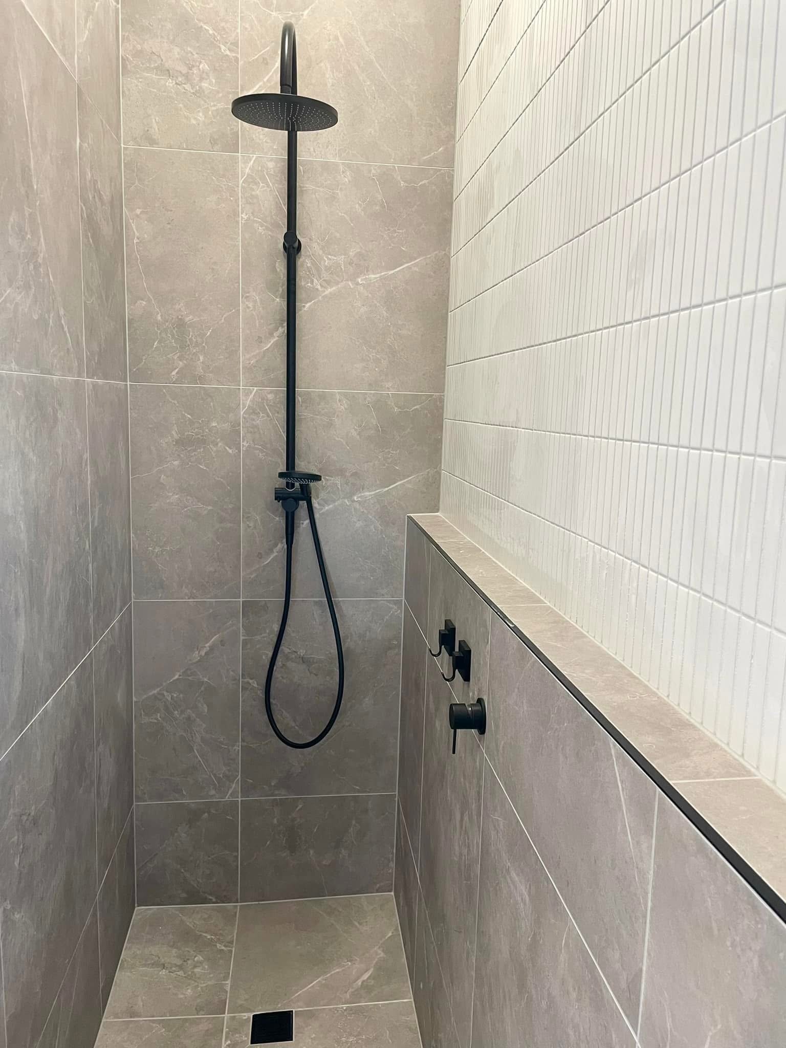 A modern shower area with gray tile walls and a ceiling-mounted black rainfall showerhead, with a handheld shower attached to a vertical rail and a black control knob on the wall.