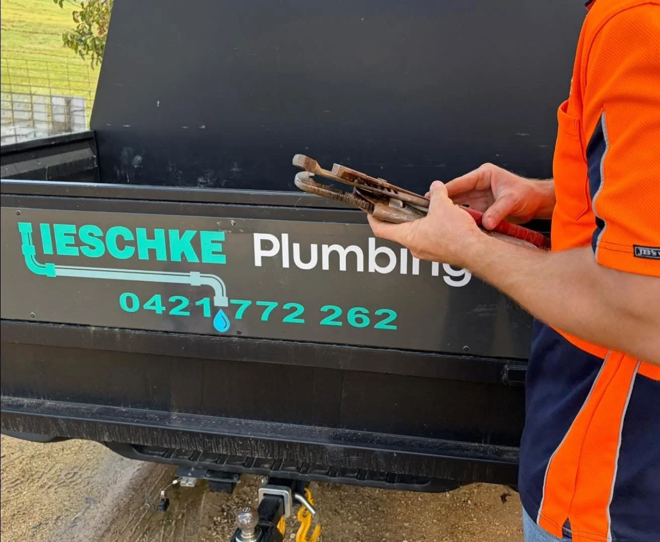 A person in an orange shirt working on a sign for a plumbing business, with a black trailer in the background displaying the business name, phone number, and plumbing pipe graphic.