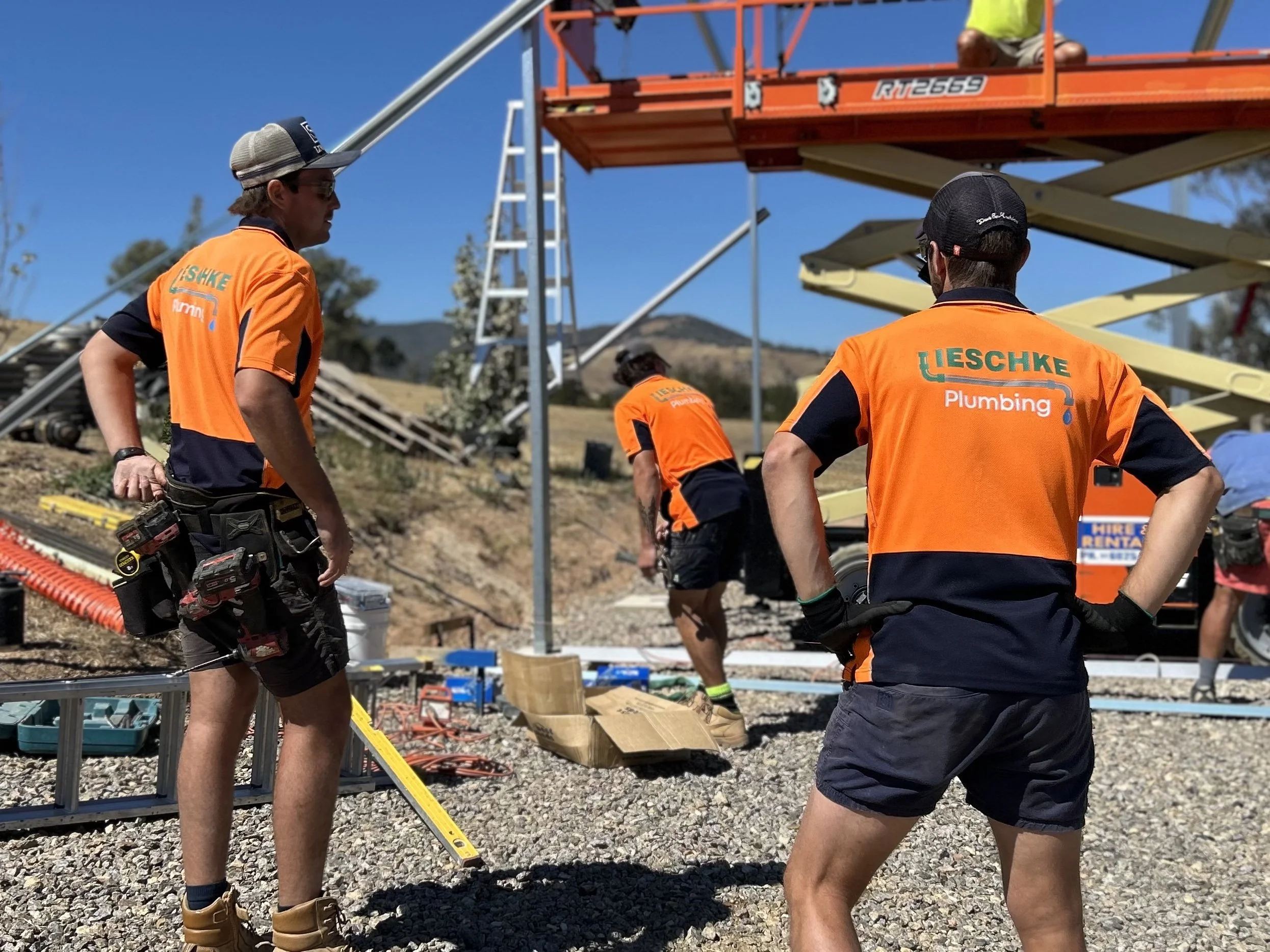Three Lieschke Plumbing workers in orange and navy hi-vis uniforms at an outdoor job site, with a scissor lift and ladder in the background.
