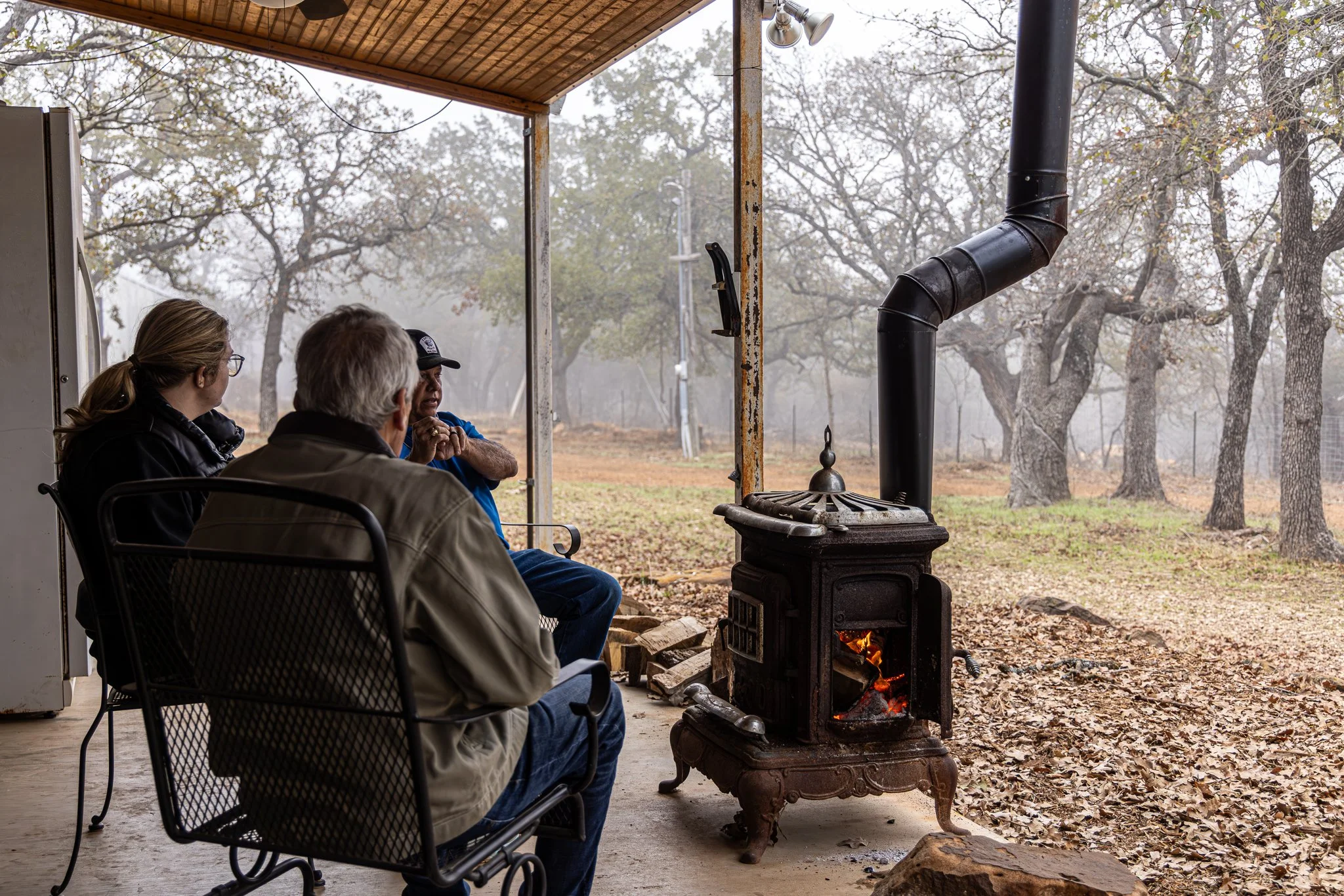 Three people sitting on chairs around an outdoor fireplace with a small fire burning, surrounded by fallen leaves and trees on a misty day.