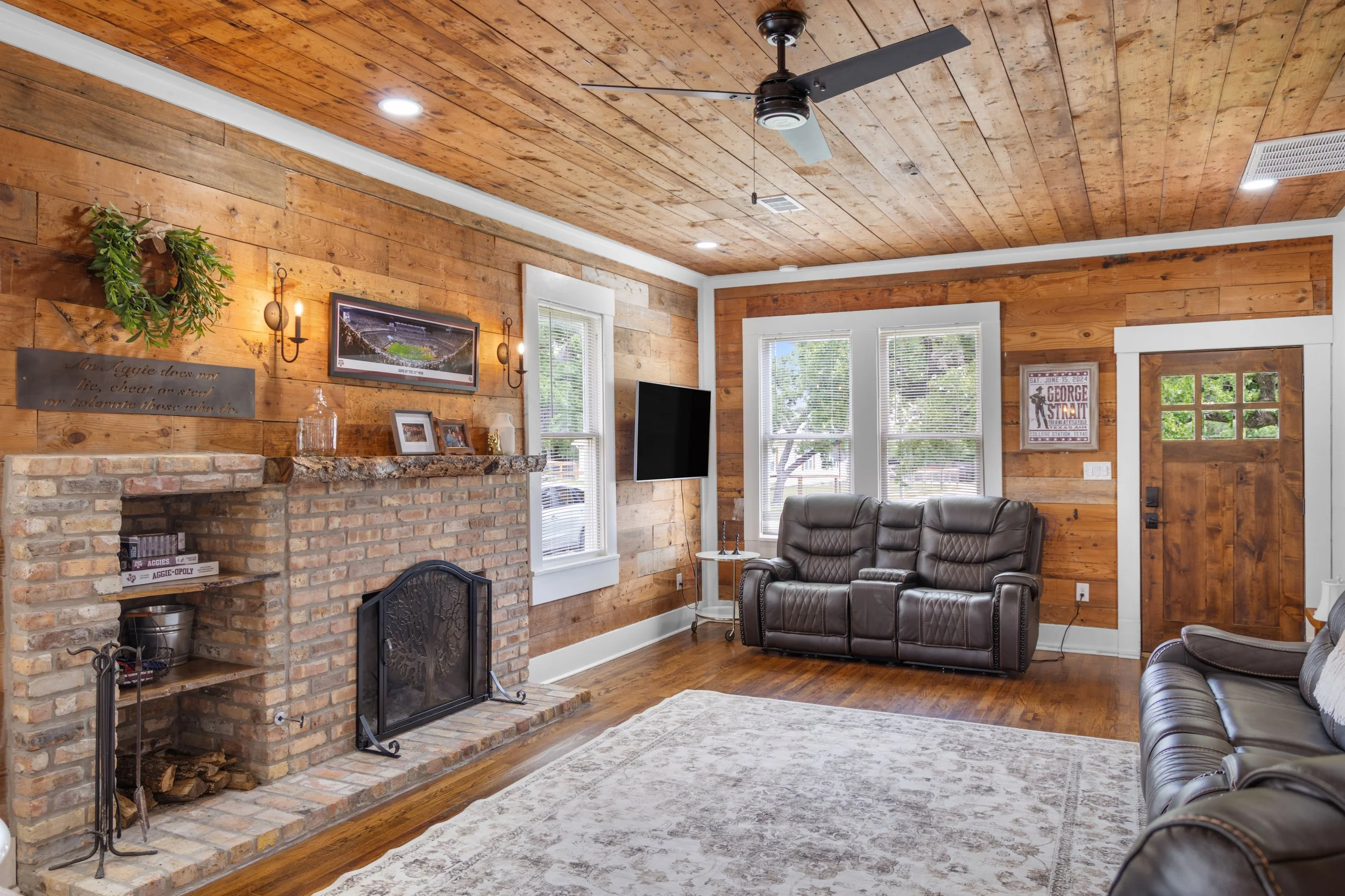 A cozy living room with wooden walls and ceiling, featuring a brick fireplace with a wooden mantel, a mounted TV, and a leather armchair. Large windows with blinds let in natural light, and framed posters and decorative plants adorn the space.