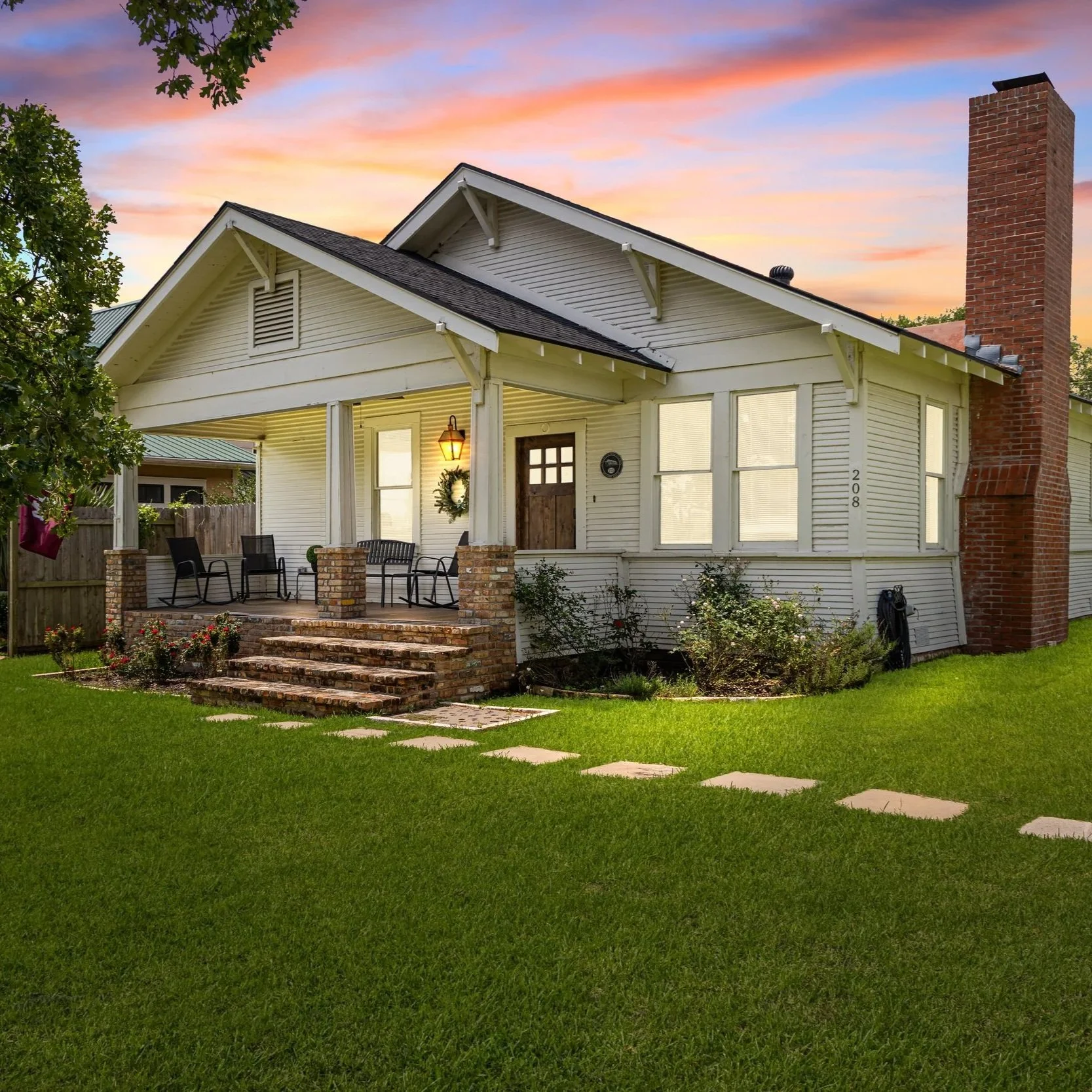 A white house with a brick chimney and a small front porch with seating, surrounded by a green lawn and stepping stones, during sunset with a colorful sky.