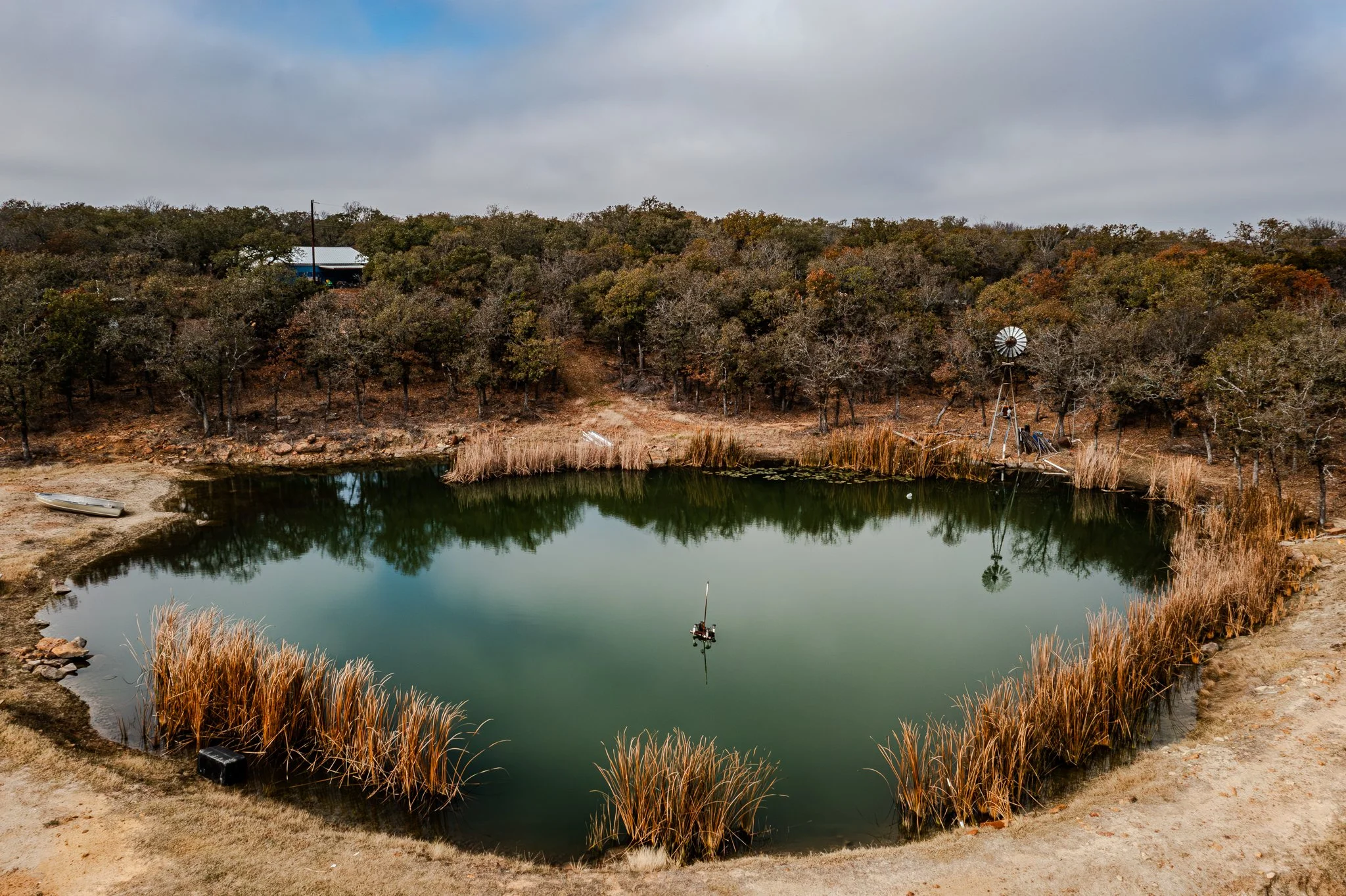 A small pond surrounded by brown grasses and trees with orange and brown leaves on a cloudy day. There is a boat pulled up on the shore, a windmill near the pond, and a small building with a metal roof on the hill in the background.