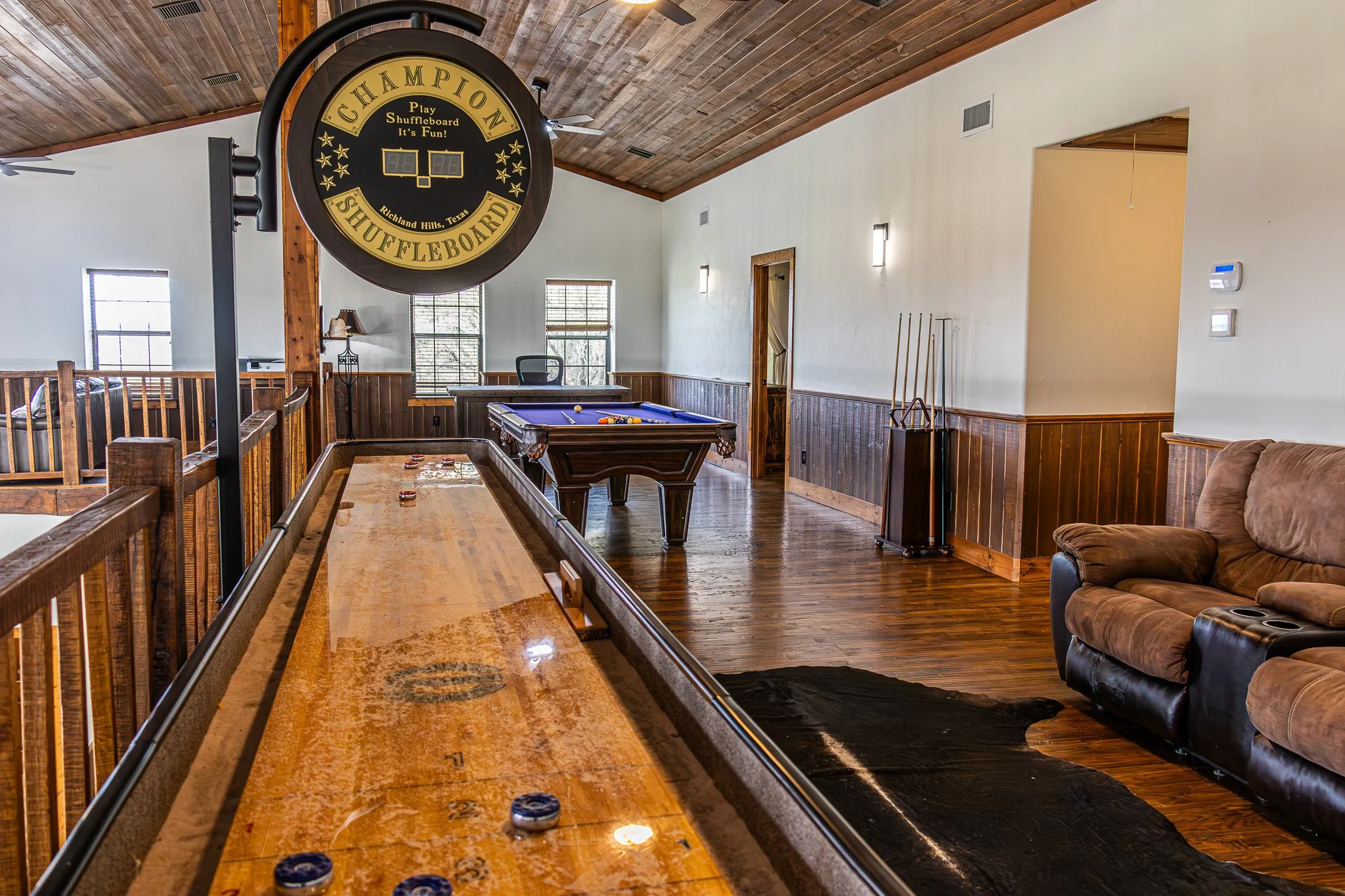 Indoor recreational room with a shuffleboard table, a pool table, a brown leather sofa, and a large circular sign that says 'Champion Shuffleboard.' The room has wooden flooring, paneled walls, and windows.