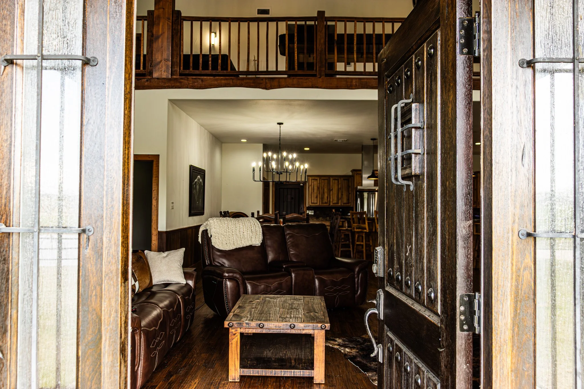 Open rustic wooden door revealing a living room with leather chairs, a wooden coffee table, wall art, and a chandelier.