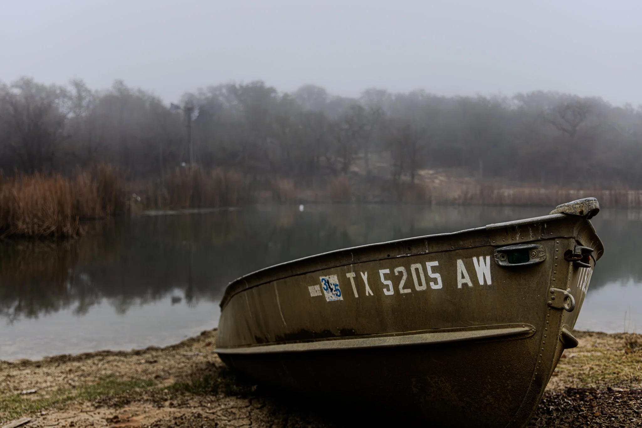 A boat resting on the shore near a calm body of water, with trees and a foggy sky in the background.