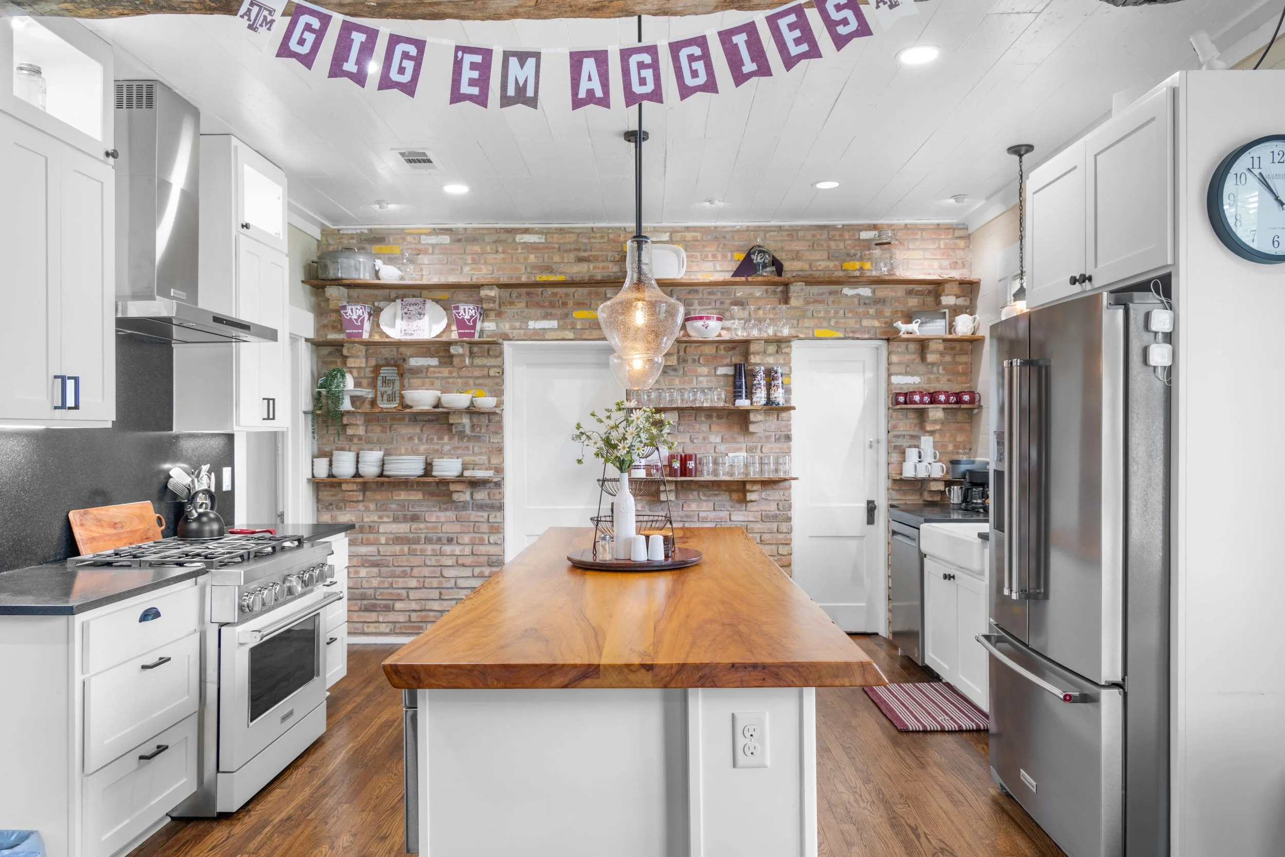 Kitchen with white cabinets and stainless steel appliances, a brick wall with wooden shelves holding dishes and decor, a wooden kitchen island with a centerpiece, and a banner that says 'Big Em Aggies' hanging from the ceiling.