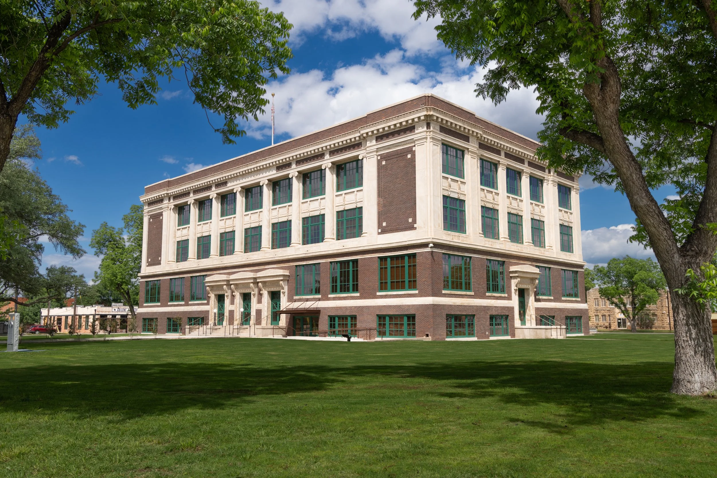 A historic brick building with large windows, surrounded by trees and green grass under a partly cloudy sky.