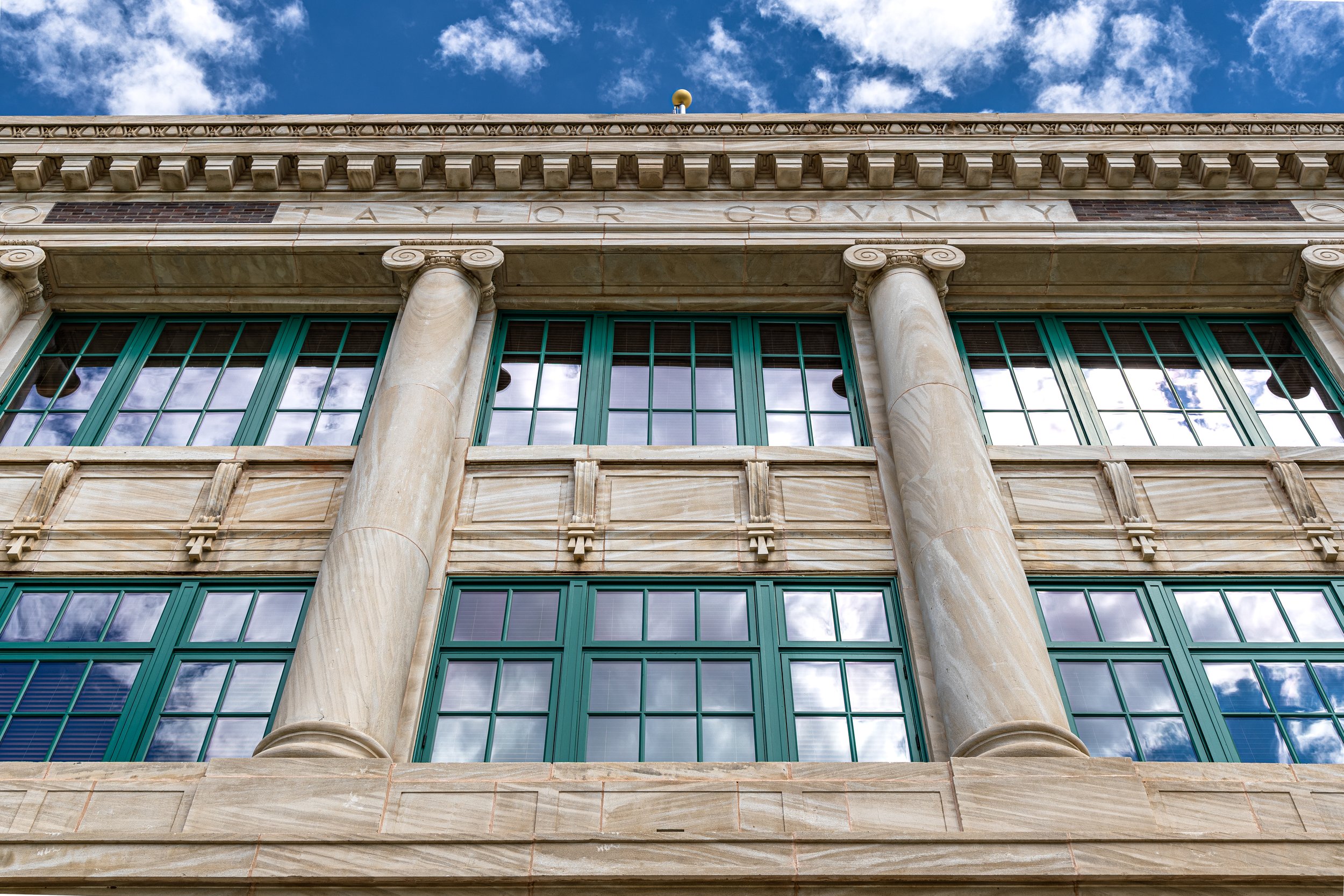Close-up of a historic courthouse building with stone columns, large green-framed windows, and a carved stone facade against a blue sky with scattered clouds.
