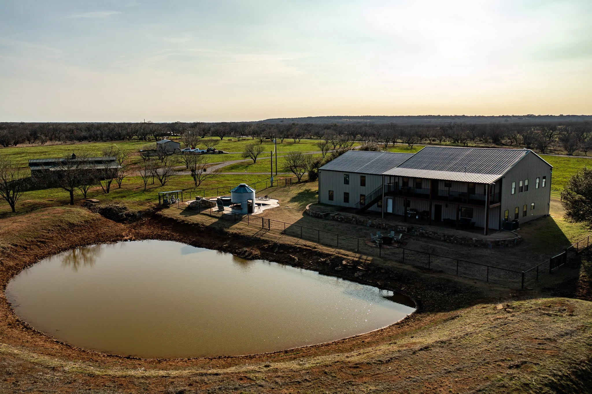 A rural farm scene at sunset showing a pond in the foreground, a large white building, smaller structures, a fenced yard, and a landscape with trees and hills in the background.