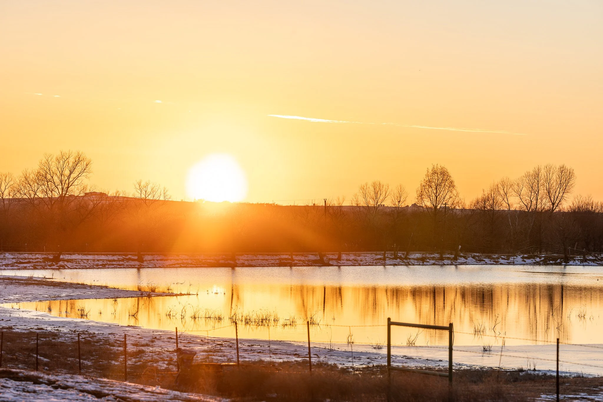 Sun setting over a frozen river with snow-covered banks, leafless trees, and a partly cloudy sky with a contrail.