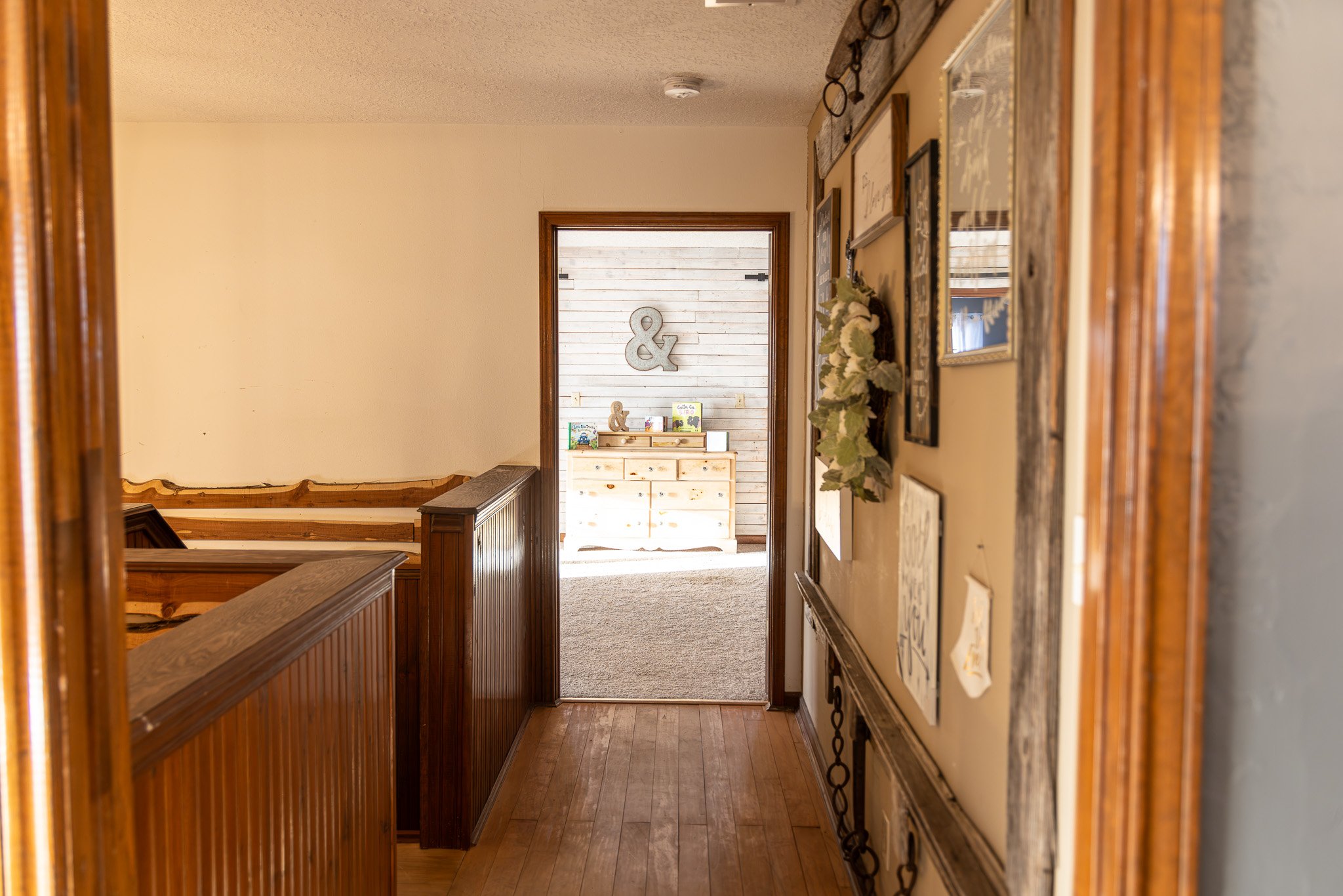 View of a cozy hallway leading into a sunlit room with decorative initials '&' on the wall, framed art, a small bookshelf with books, and a flower arrangement.