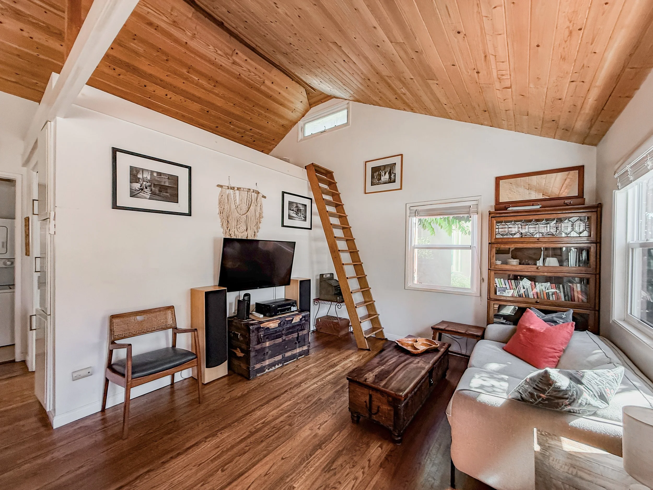 Living room with wooden ceiling, hardwood floors, a TV on a stand, framed photos on the wall, a ladder leading to a loft, a window with blinds, a wooden cabinet with glass doors, a beige sofa with cushions, a wooden coffee table, and a wicker chair.