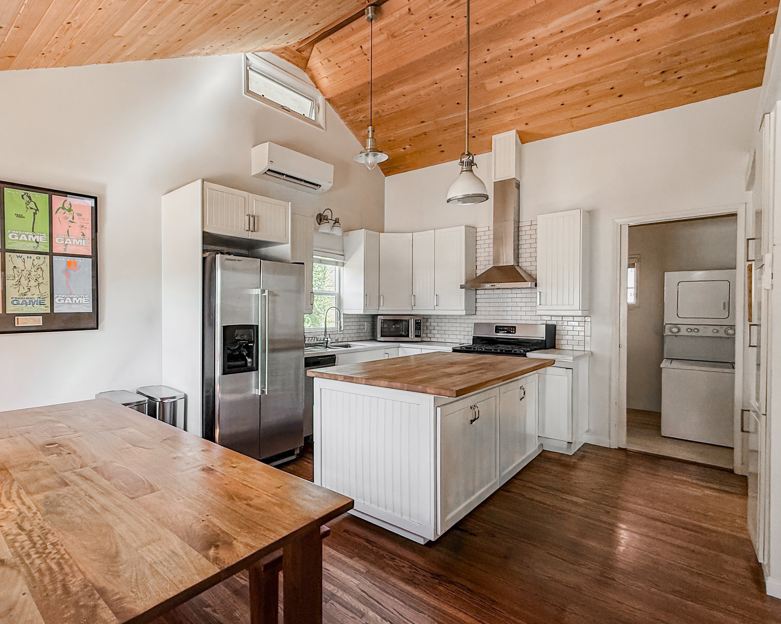 Interior view of a kitchen with a wooden ceiling, white cabinets, a stainless steel refrigerator, microwave, oven, and washer/dryer. There are pendant lights and a window above the sink.