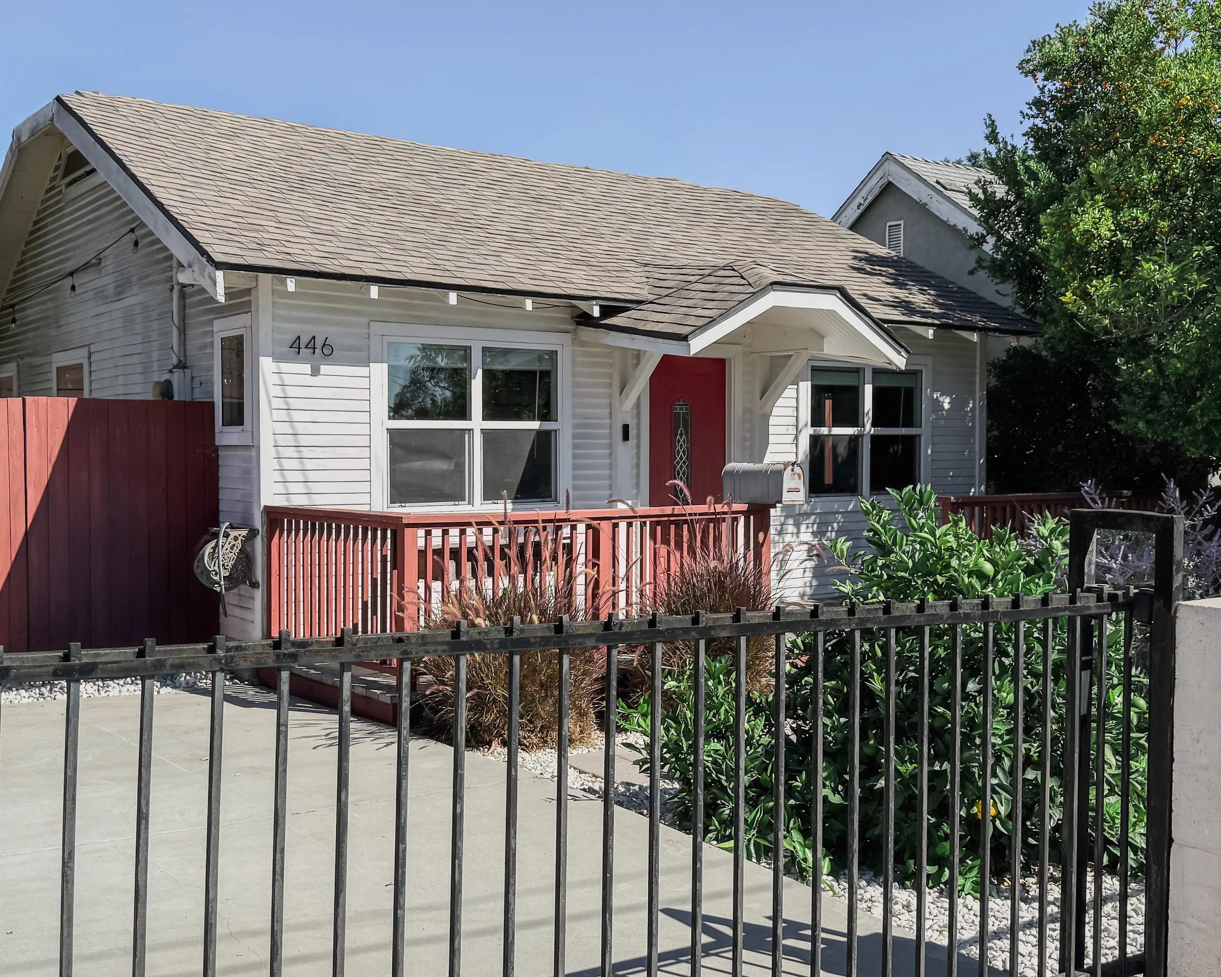 A white house with the number 446 on the wall, a red front door, and a small front porch, surrounded by a black metal fence, with a tree and greenery nearby and a clear blue sky.