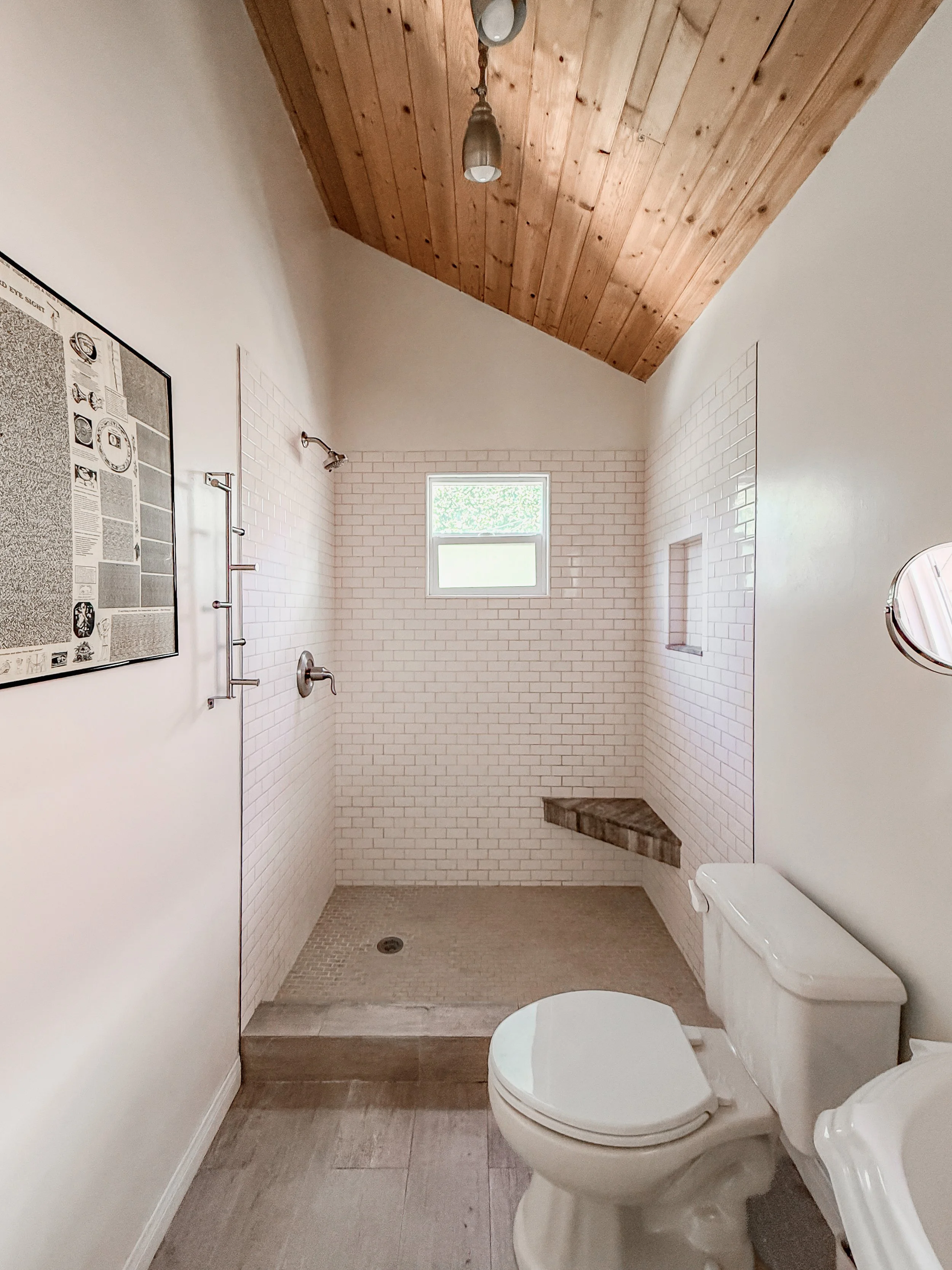 A modern bathroom with a wooden ceiling, white subway tile walls, a small window, a walk-in shower with a shower head and a built-in bench, a toilet, and a mirror.