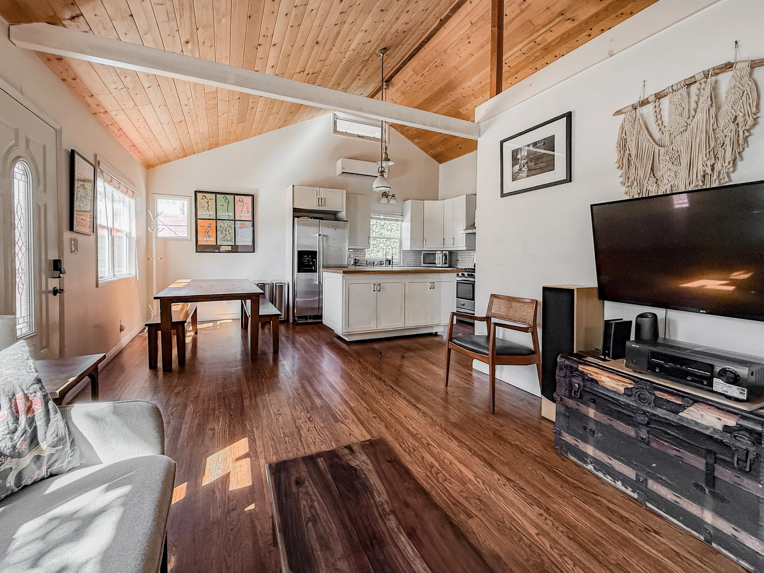 Living room with a sloped wooden ceiling, a framed poster on the wall, a gray sofa with pillows, a dining table, a kitchen with white cabinets, and a mounted flat screen TV.
