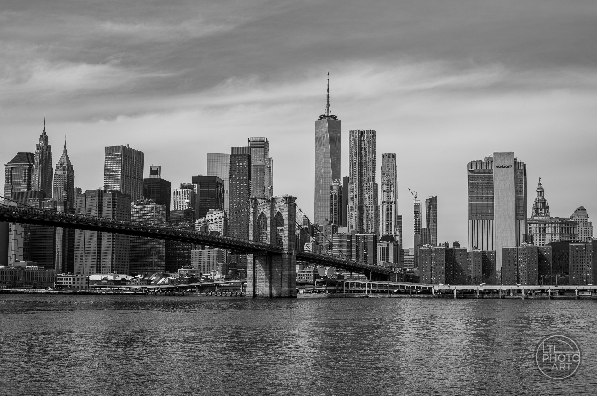 Black-and-white view of the Brooklyn Bridge crossing the East River toward the Lower Manhattan skyline and One World Trade Center.