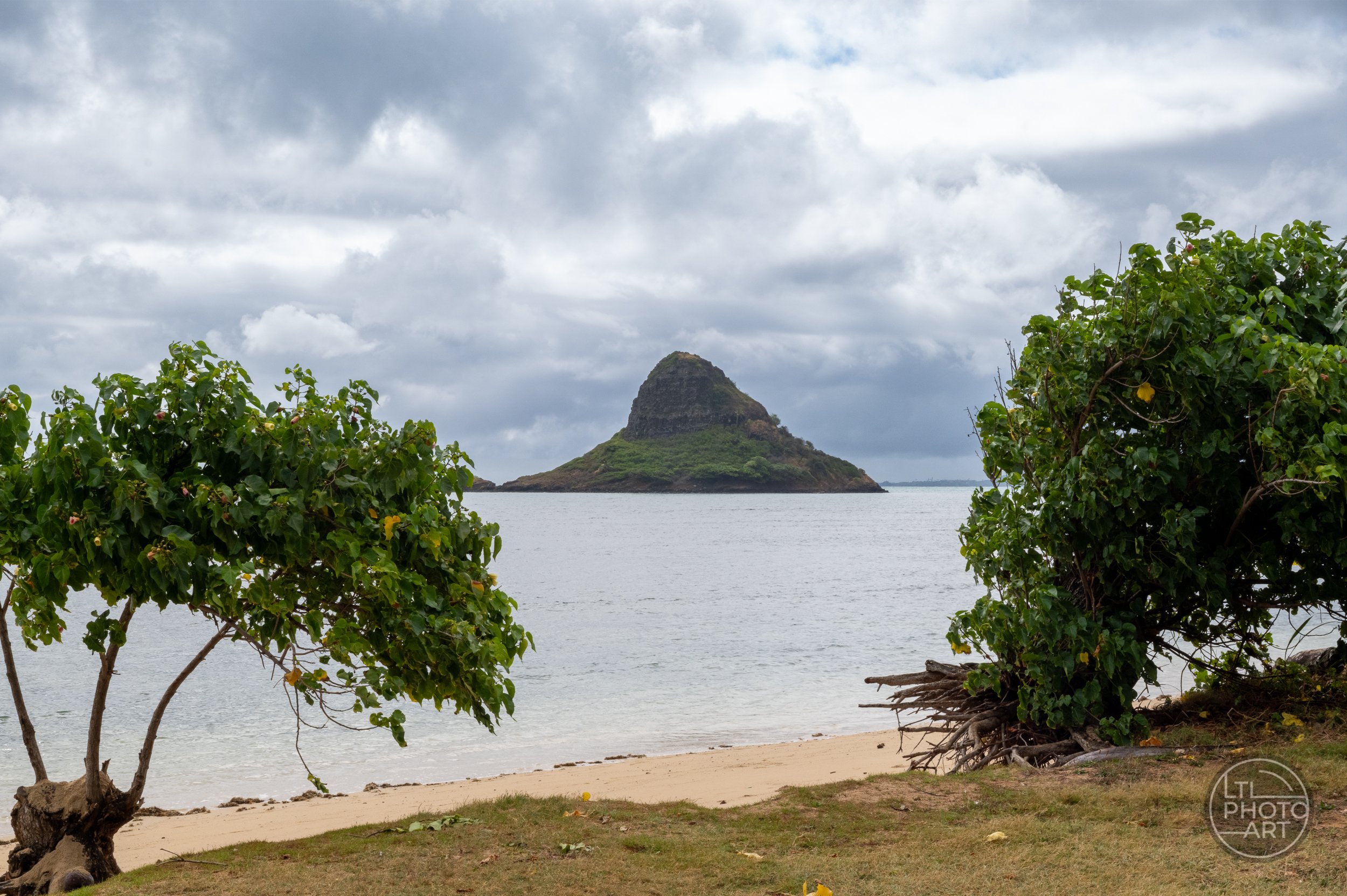 Mokoliʻi island off the windward coast of Oahu, Hawaii, viewed across calm water and framed by tropical trees beneath cloudy skies.