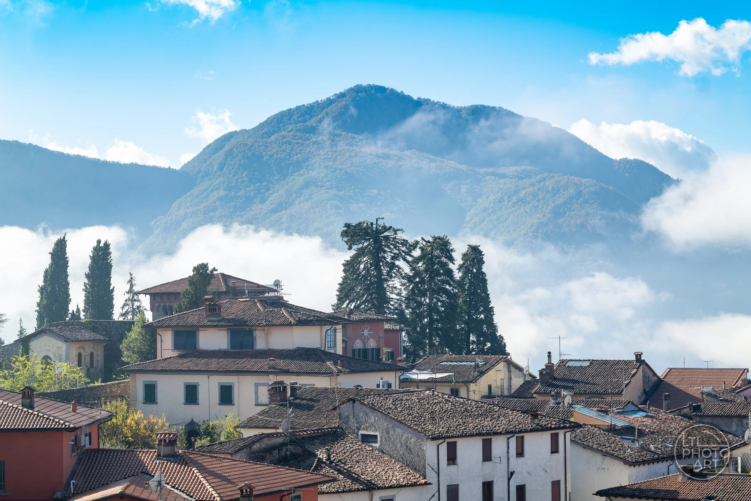 Historic tiled rooftops in Barga, Tuscany, with trees, drifting clouds, and the Apuan Alps rising in the background.