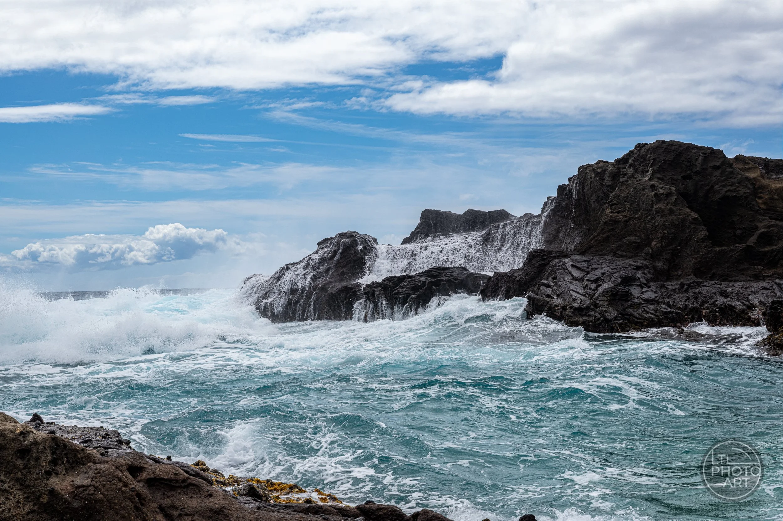 Ocean waves spill across dark volcanic rocks at Halona Beach Cove on Oahu beneath a blue sky.