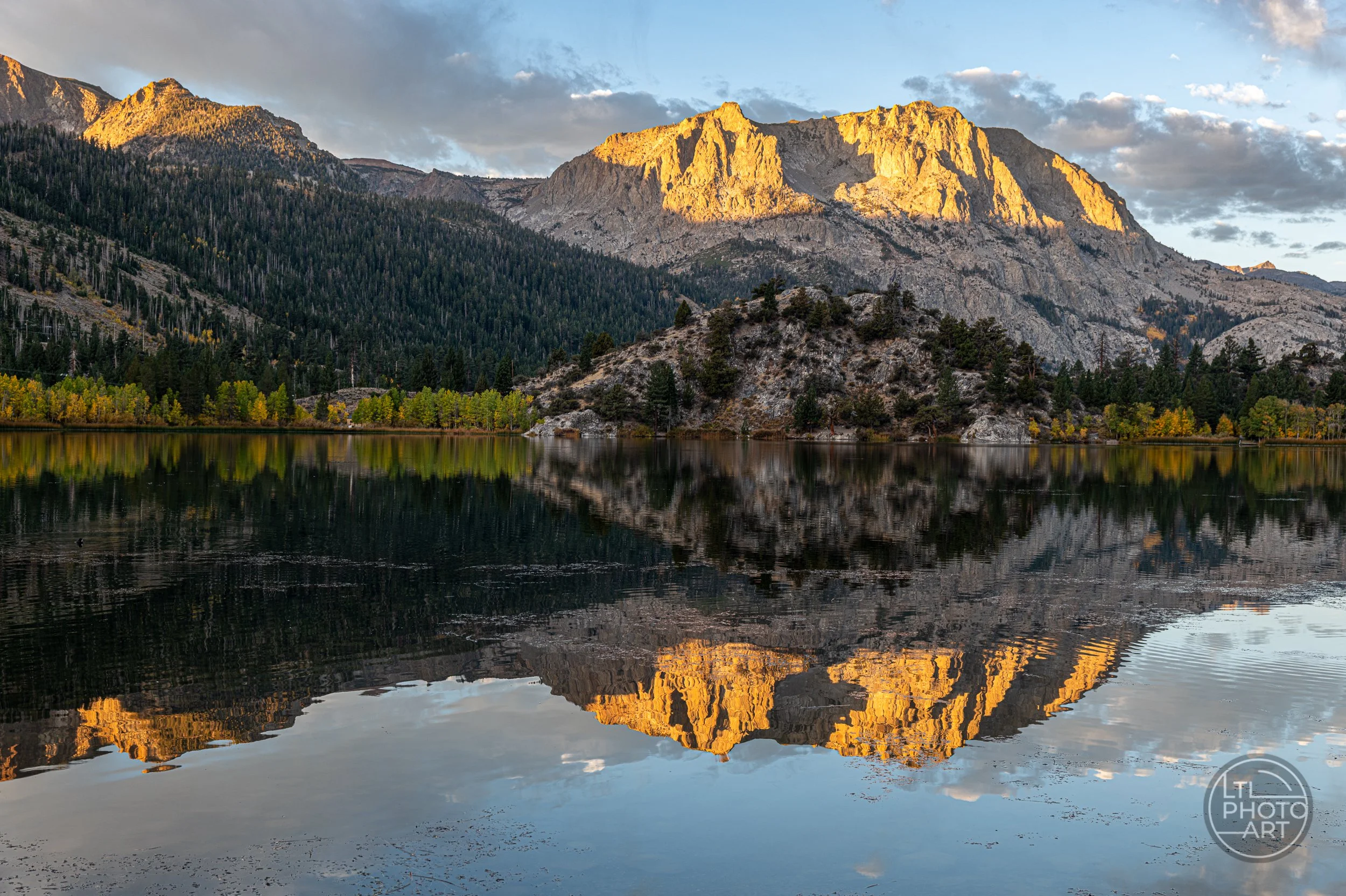 Landscape photograph of Gull Lake in California's June Lake Loop, featuring granite mountain peaks illuminated by golden evening sunlight, evergreen forests, autumn foliage, and reflections in the calm lake water.